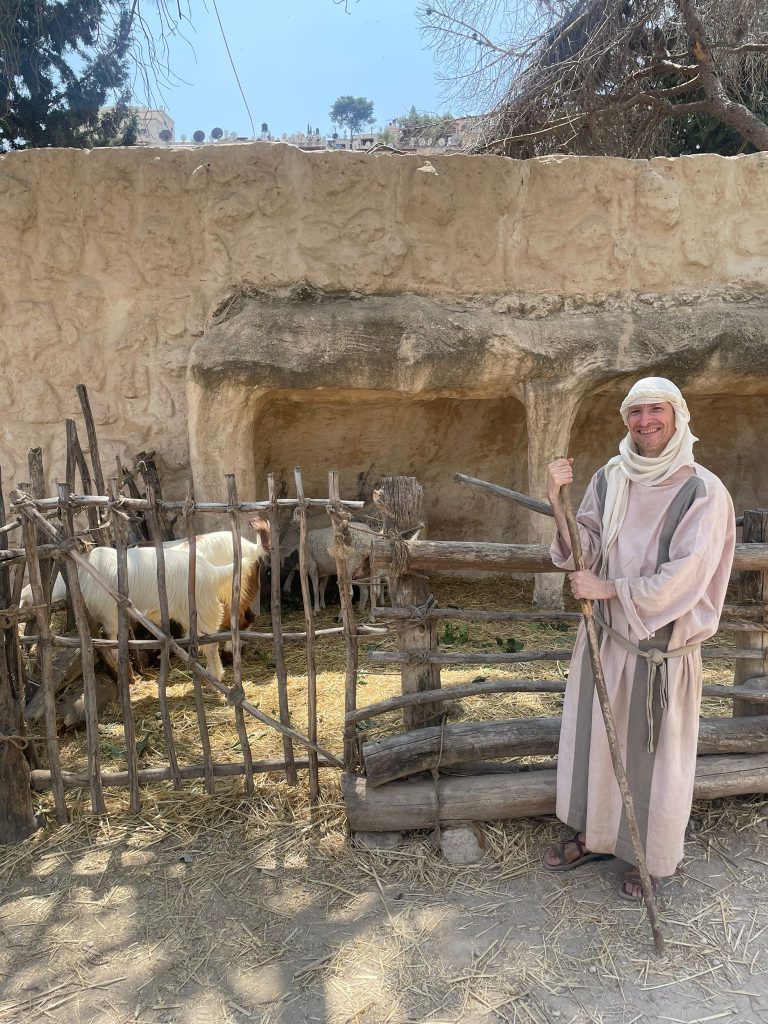 A photo of Brian Murray dressed up in 1st-Century clothing standing in front of the sheepfold at the Nazareth Village.